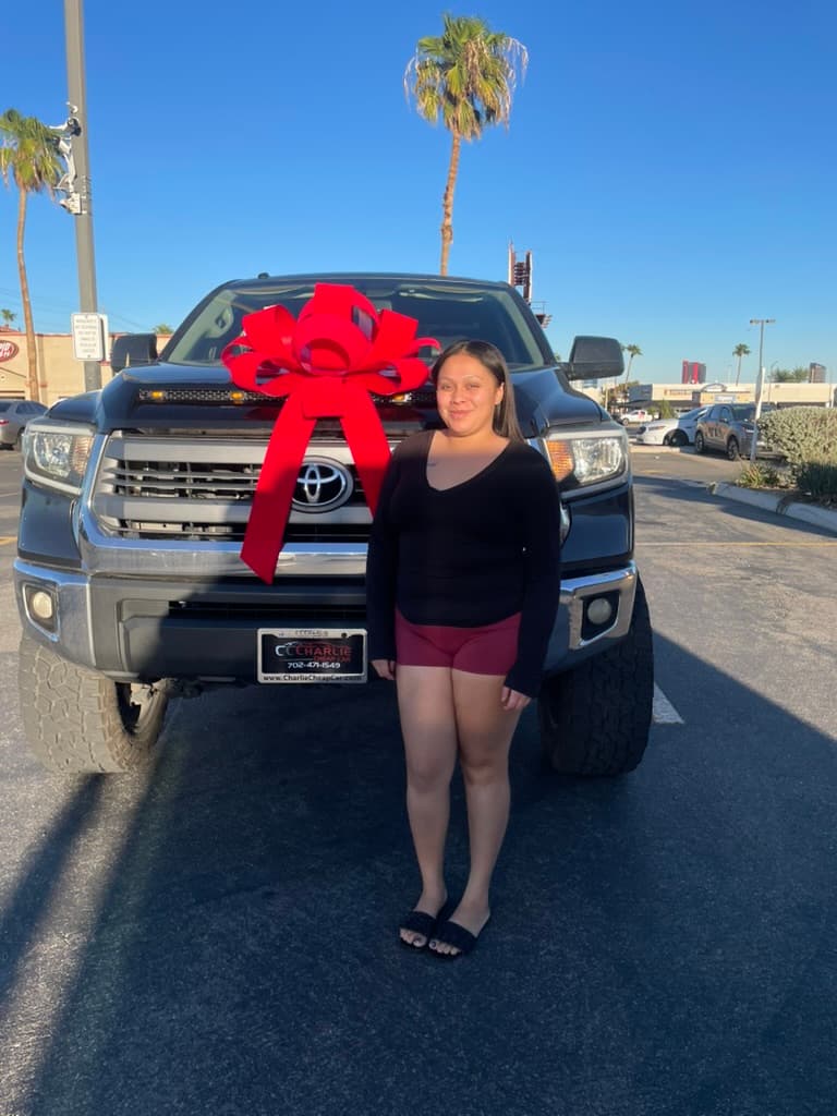 A smiling customer standing in front of her black Toyota Tundra pickup with a large red bow on the grille, on the Charlie Cheap Car lot