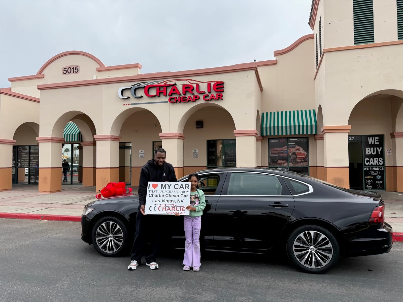 An adult and a child holding the I love my car sign beside their black Volvo S90 with a red bow, outside the Charlie Cheap Car storefront