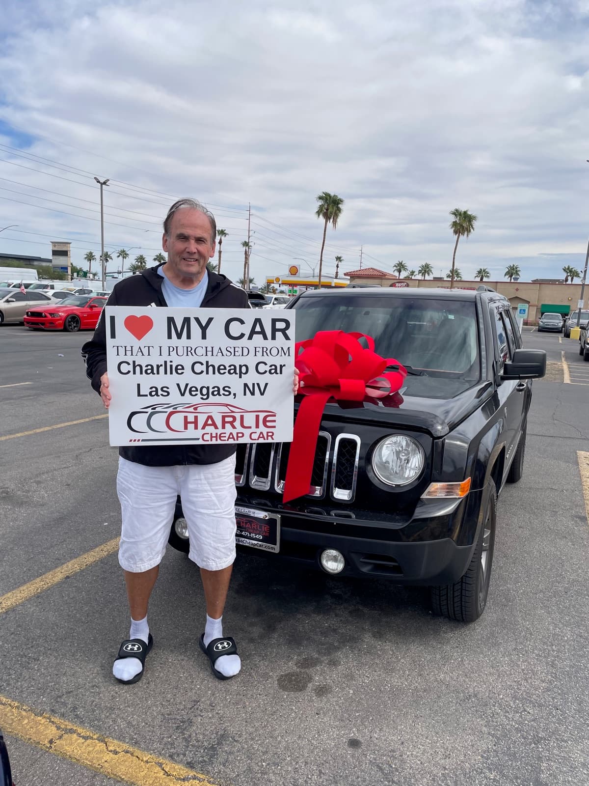 A smiling customer holding the I love my car sign beside their black Jeep Patriot with a big red bow, on the Charlie Cheap Car lot