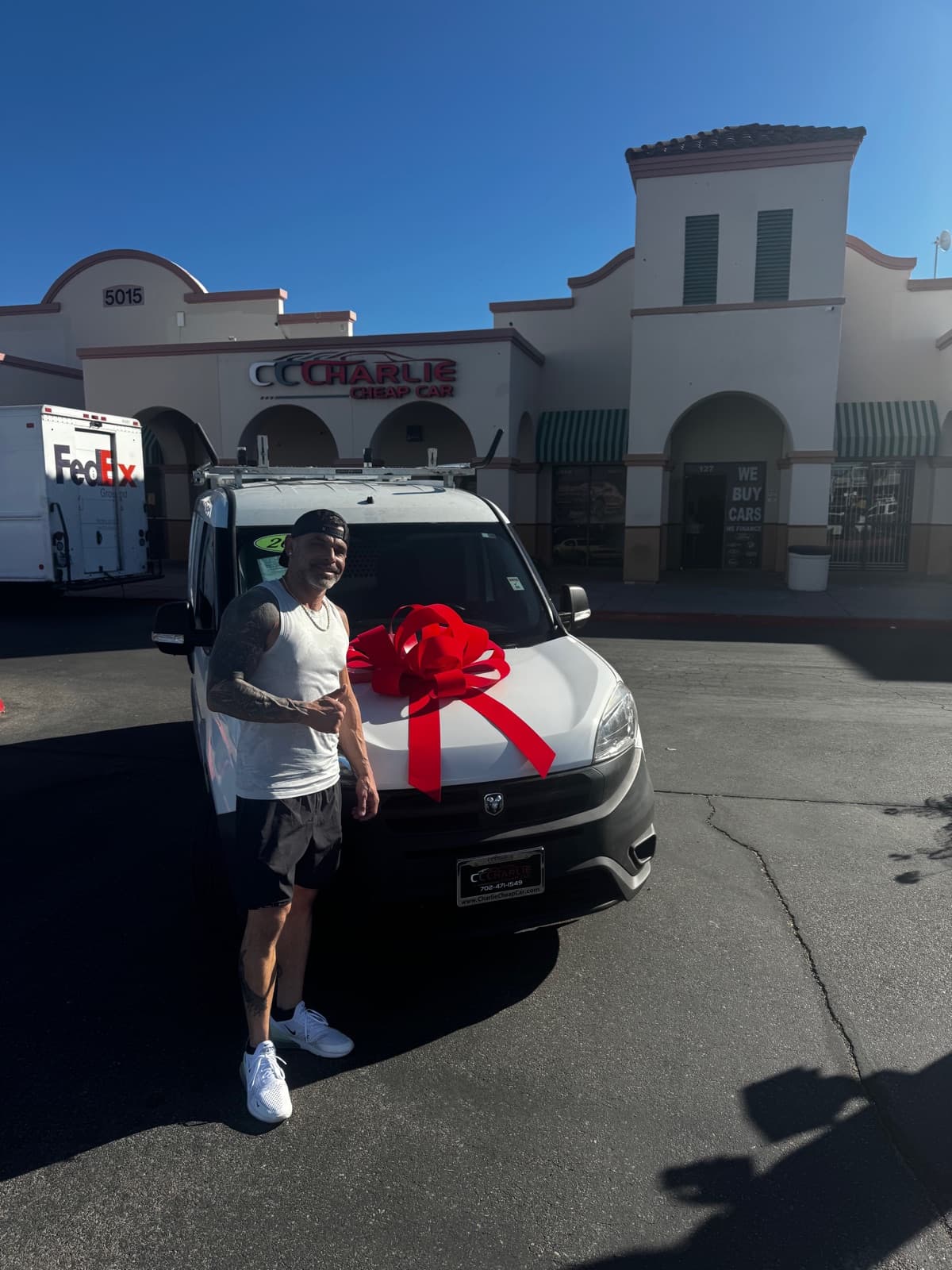 A smiling customer giving a thumbs up beside their white Ram ProMaster City van with a big red bow, outside the Charlie Cheap Car storefront