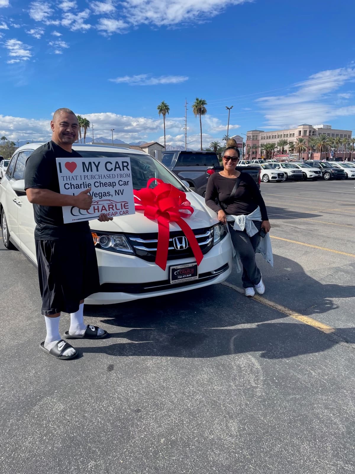 Two smiling customers, one holding the I love my car sign, beside their white Honda Odyssey minivan with a big red bow