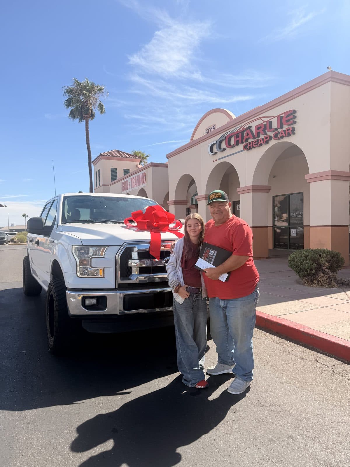 A father and daughter beside their white Ford F-150 with a red bow, outside Charlie Cheap Car