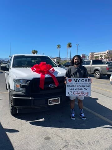 A customer holding the I love my car sign beside his white Ford F-150 pickup with a big red bow on the hood, on the Charlie Cheap Car lot
