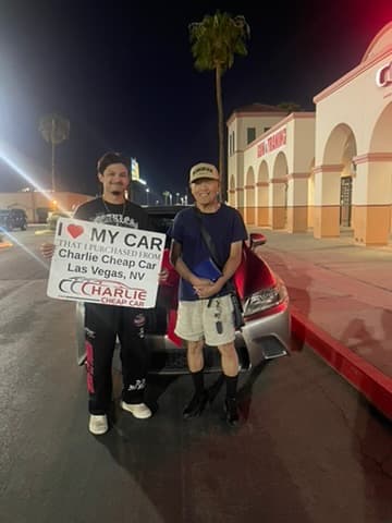Two customers at night outside the Charlie Cheap Car storefront, one holding the I love my car sign, in front of their red sedan