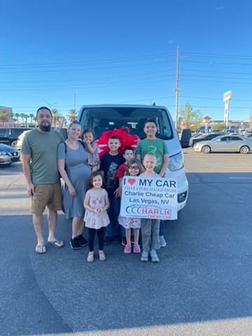 A family of two parents and seven children holding the I love my car sign in front of their white passenger van with a big red bow, on the Charlie Cheap Car lot