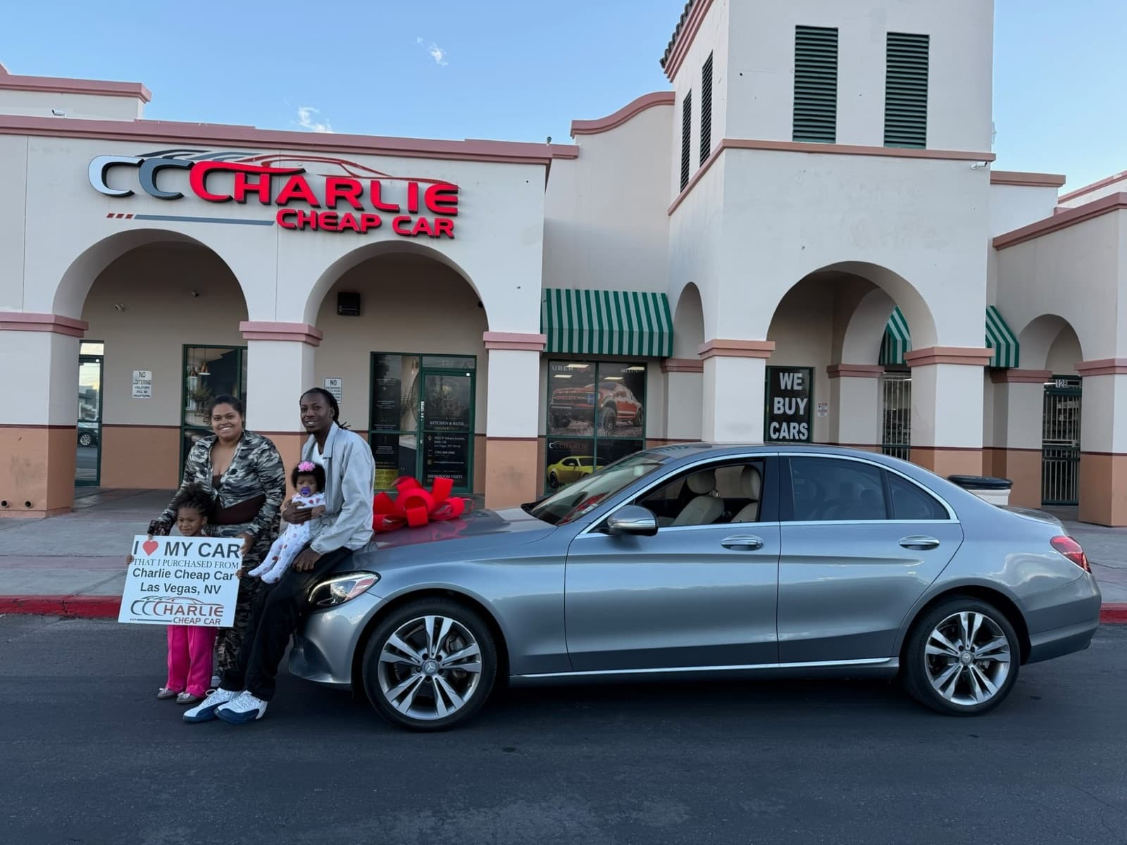 A young family with two little girls and the I love my car sign beside their silver Mercedes-Benz C-Class with a big red bow, outside the Charlie Cheap Car storefront
