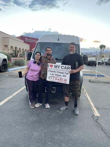 Three customers holding the I love my car sign in front of their white high-roof van on the Charlie Cheap Car lot
