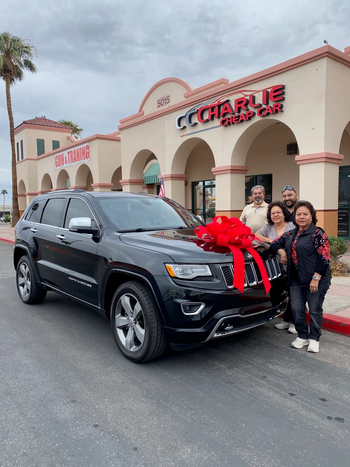 Four customers beside their black Jeep Grand Cherokee with a big red bow on the hood, outside the Charlie Cheap Car storefront