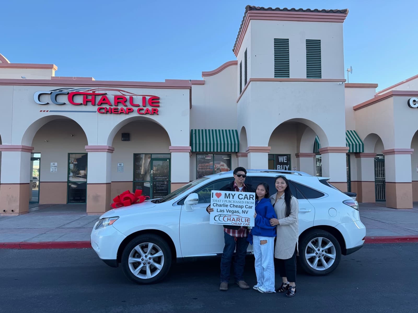 A family of three holding the I love my car sign beside their white Lexus RX with a big red bow, outside the Charlie Cheap Car storefront