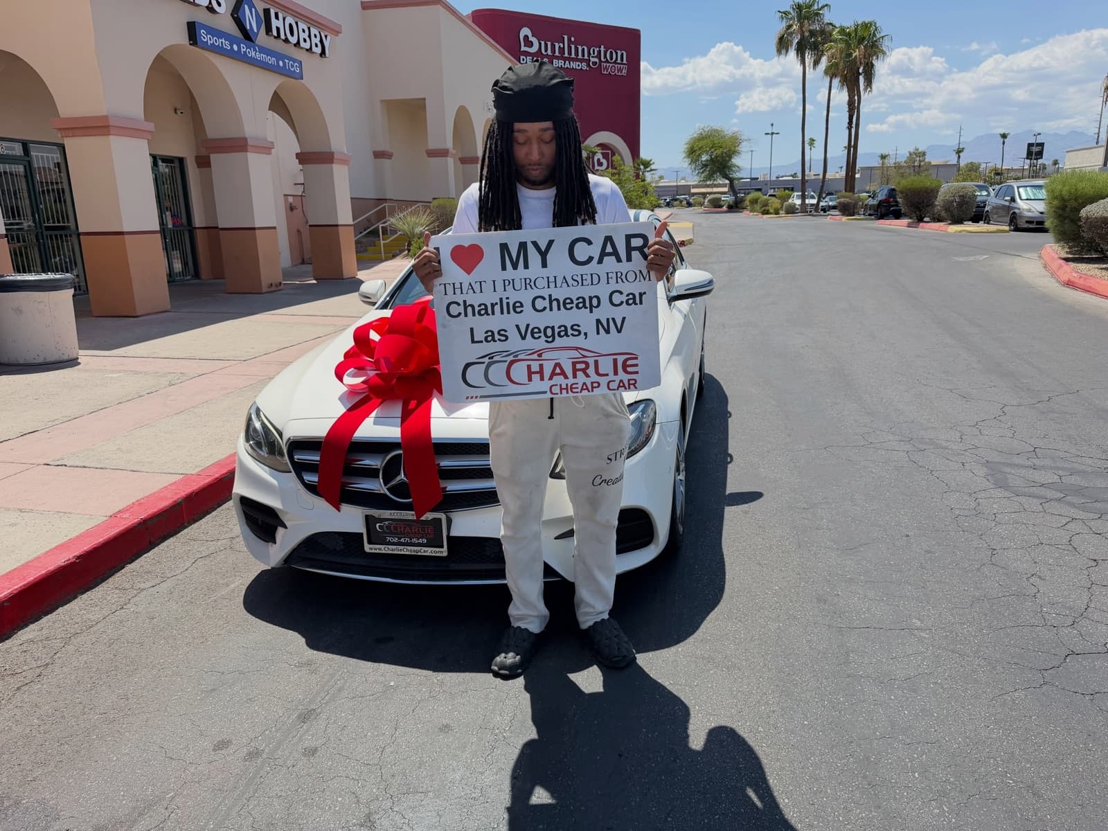 A customer holding the I love my car sign in front of a white Mercedes-Benz sedan with a red bow, outside Charlie Cheap Car