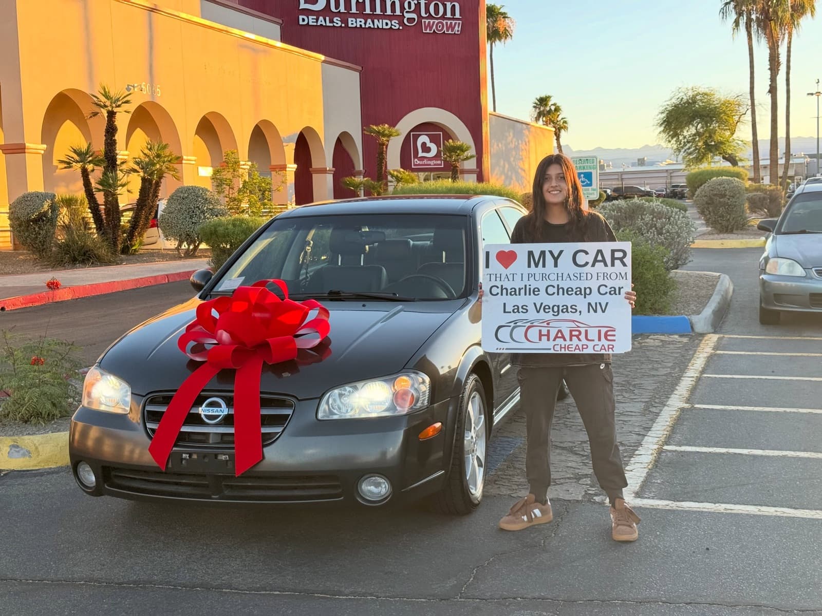 Customer holds an I love my car sign beside a grey Nissan Maxima with a red bow