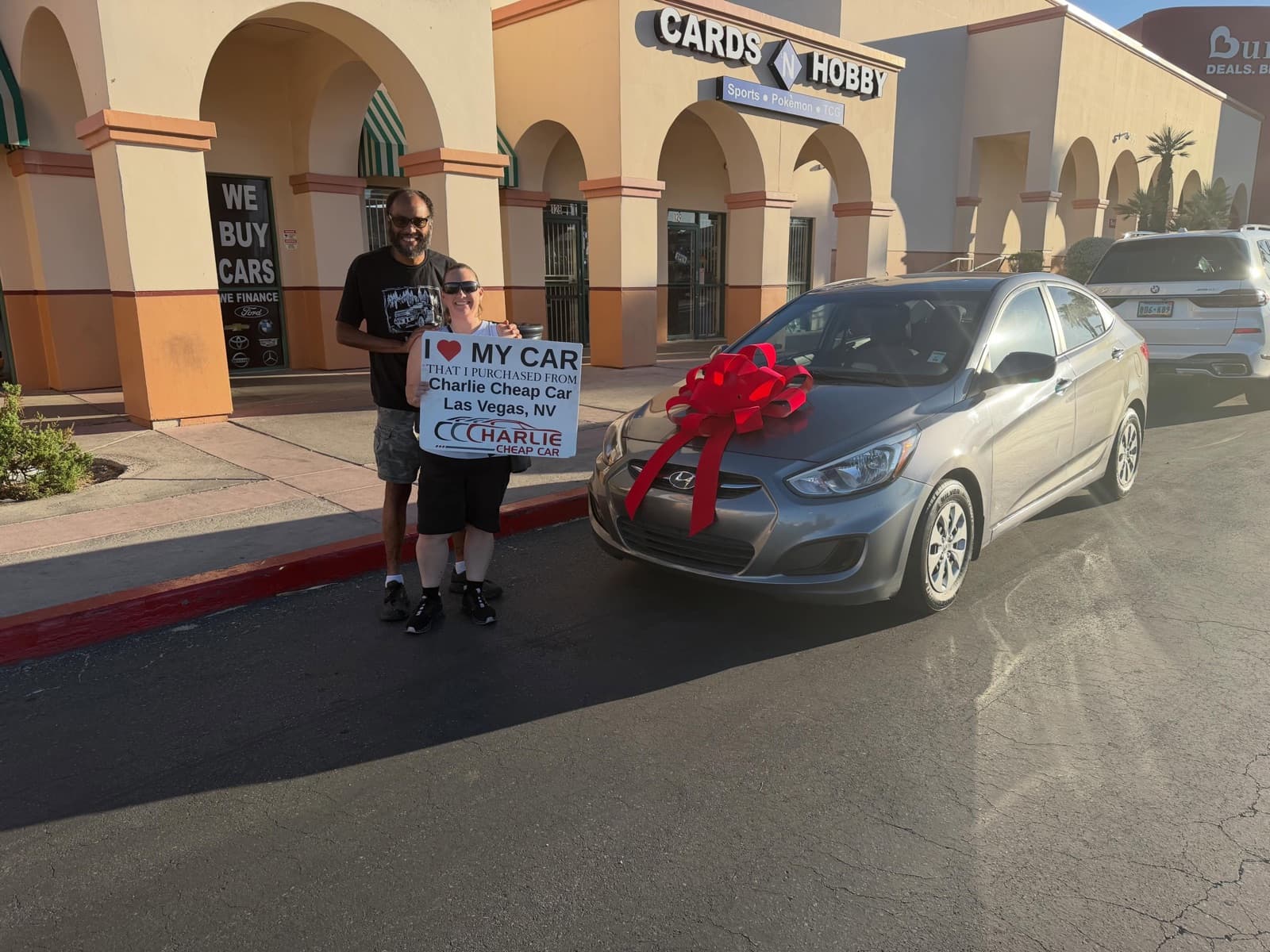 Two customers holding an I love my car sign beside a gray Hyundai Accent with a red bow