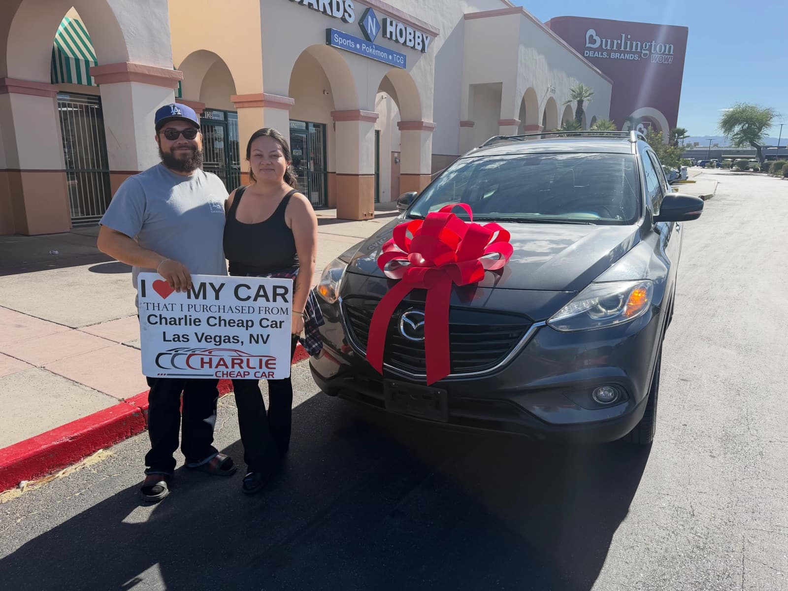 Two customers holding the I love my car sign beside a dark gray Mazda CX-9 with a red bow