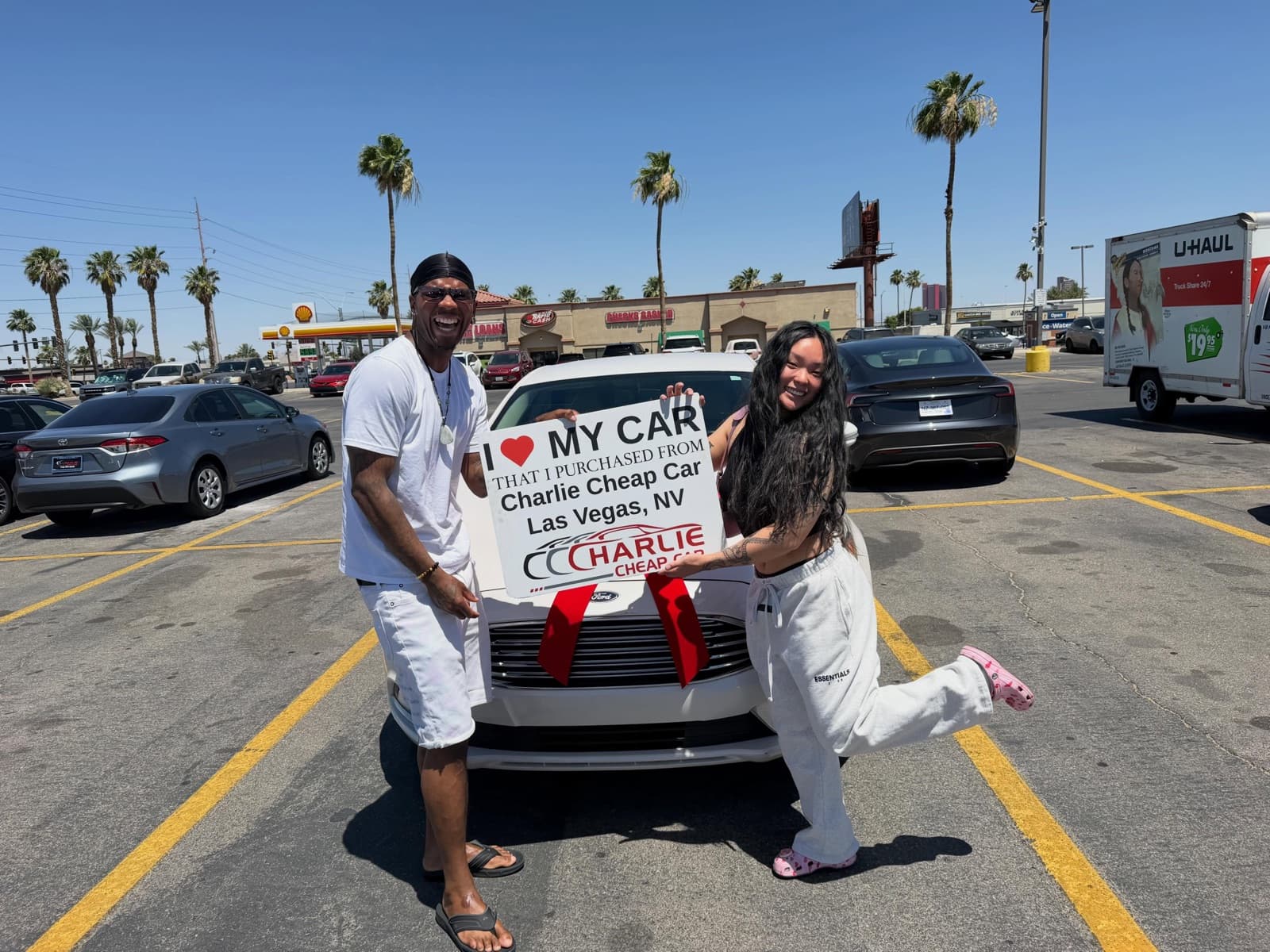 Two customers laughing while holding the I love my car sign in front of a white Ford sedan