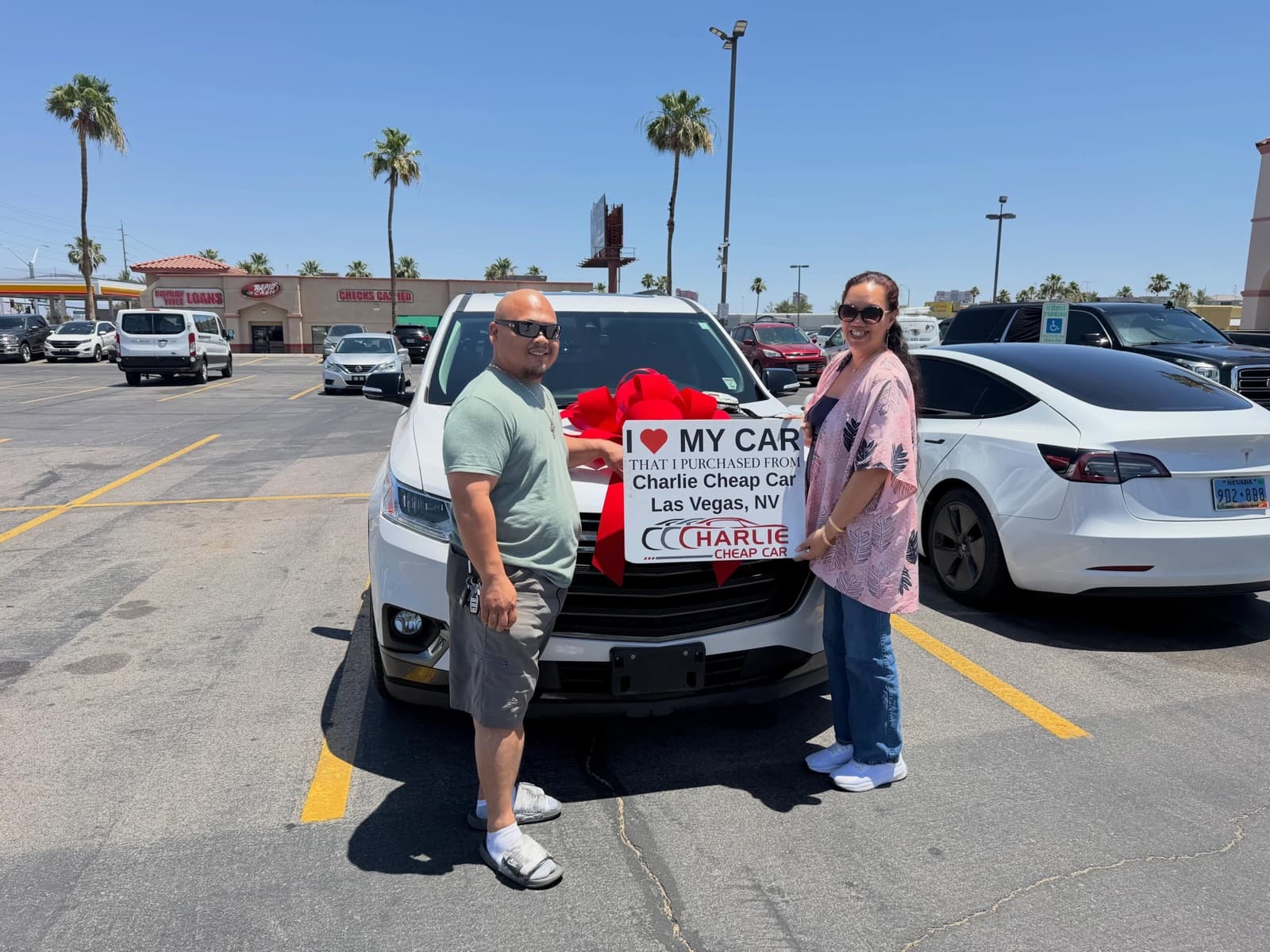Two customers holding the I love my car sign in front of a white Chevrolet Traverse with a red bow